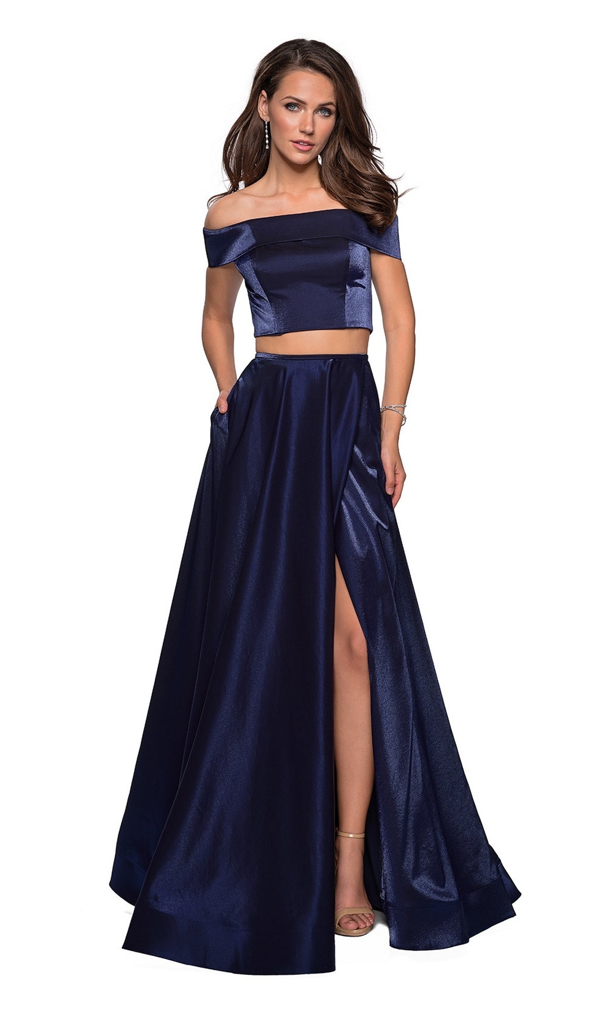 A woman poses against a white background wearing the La Femme A-Line Two-Piece Satin Prom Dress 26919, featuring a navy blue off-the-shoulder crop top and matching high-waisted, floor-length skirt with an A-line silhouette and thigh-high slit.